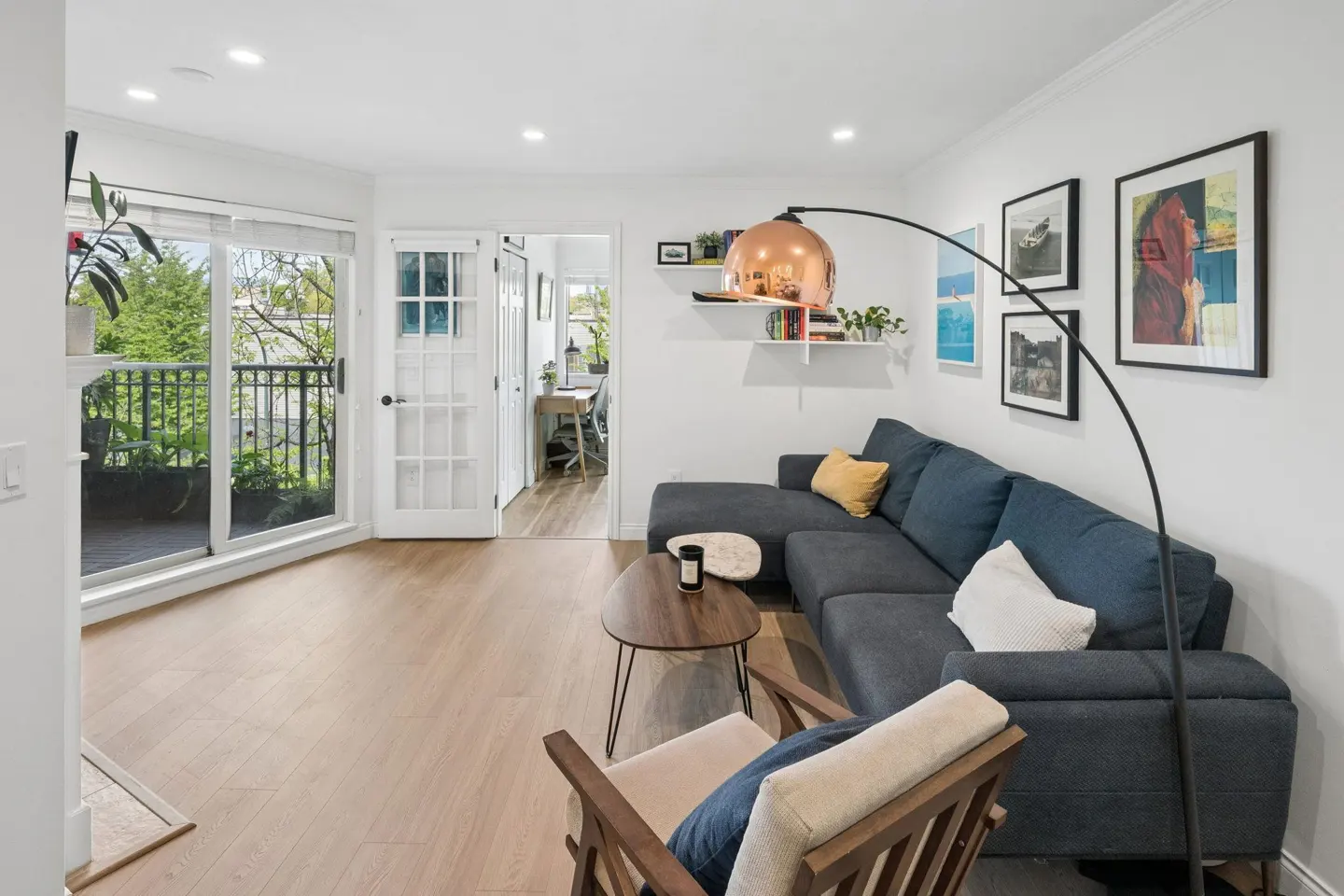 Bright living room with wood floors, a blue sectional sofa, and a copper arc lamp. Balcony with sliding glass doors and an office visible through a doorway.