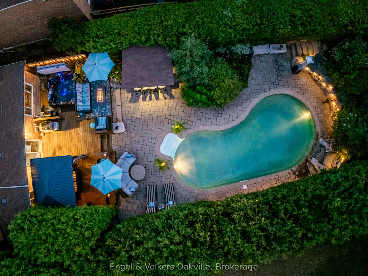 Aerial view of a backyard with a kidney-shaped pool, patio, hot tub, and lush green hedges. Outdoor furniture and lighting create an inviting atmosphere.