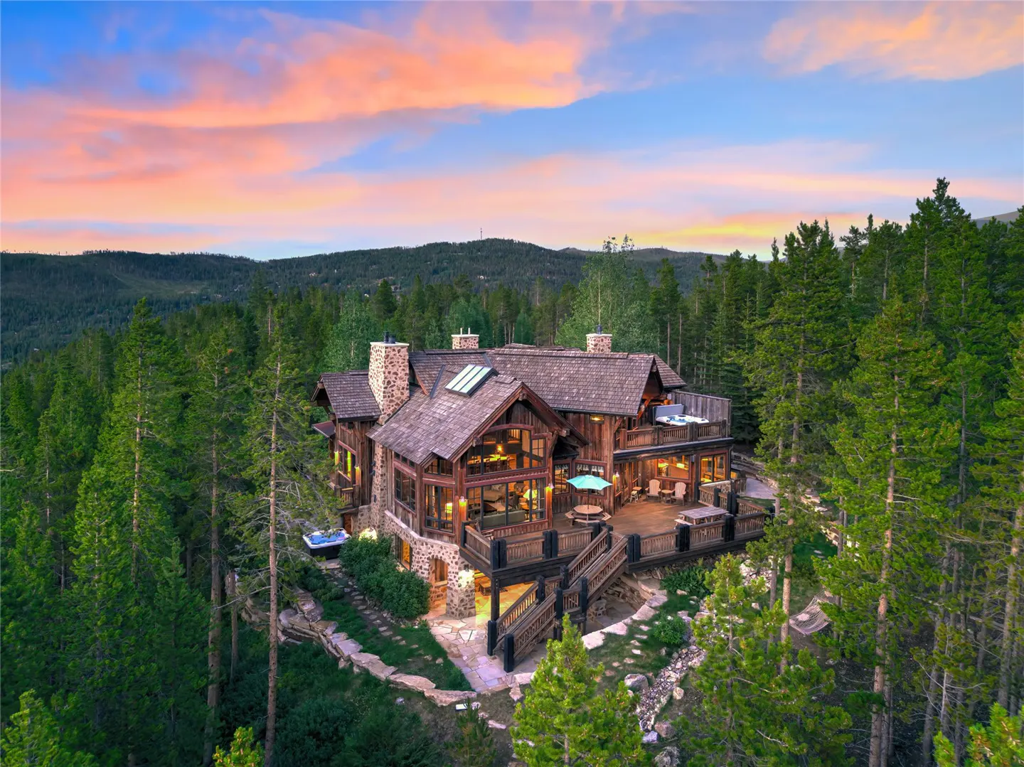 A large, brown, wood and stone house with a deck is surrounded by tall green trees under a colorful sunset sky.