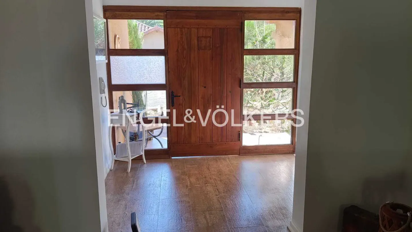 A wooden front door with glass side panels opens to a bright entryway with wood-look tile flooring and a small side table.