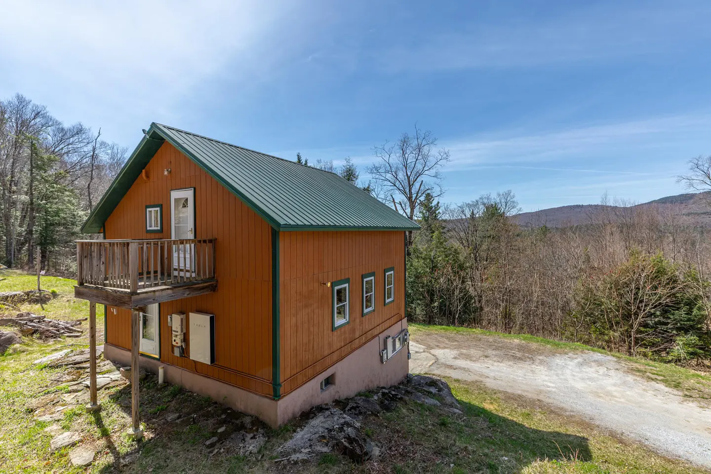 Exterior view of a two-story orange house with a green metal roof and a small wooden balcony. The house is surrounded by trees and a mountain range in the background.