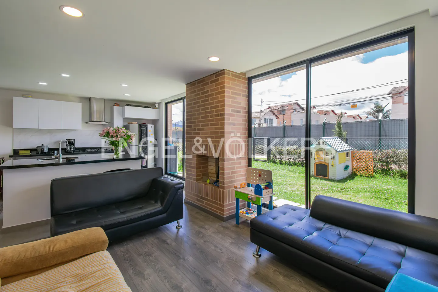 Open-concept living space with a brick fireplace, black leather sofas, and a view of a green backyard with a playhouse.