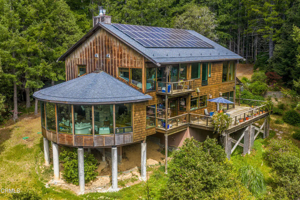 Exterior view of a wood-shingled house with solar panels, a round glass-enclosed room, and a deck, surrounded by trees.