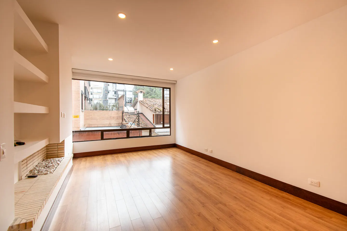 Bright, empty room with hardwood floors, white walls, and a large window showing a cityscape. Built-in shelves and a faux fireplace add architectural detail.
