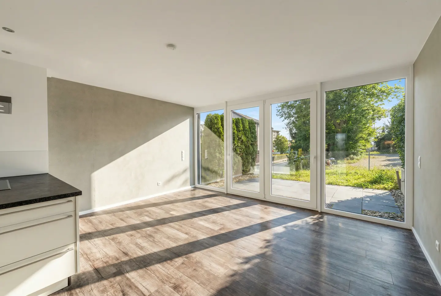 Bright, empty room with dark wood floors and gray walls. Sunlight streams through large white-framed glass doors to a green yard. Kitchen cabinets are visible.