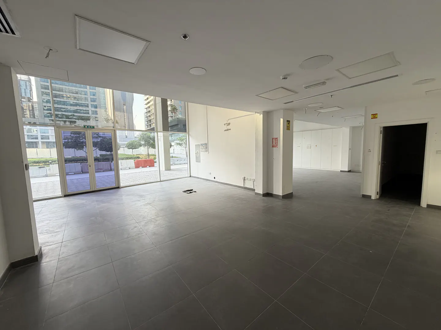 An empty commercial space with gray tile floors, white walls, and large windows showing a city view.