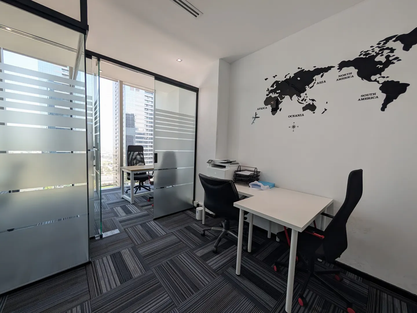 Office space with white walls, a black world map, and two desks with black chairs. Glass-walled offices overlook a city skyline.