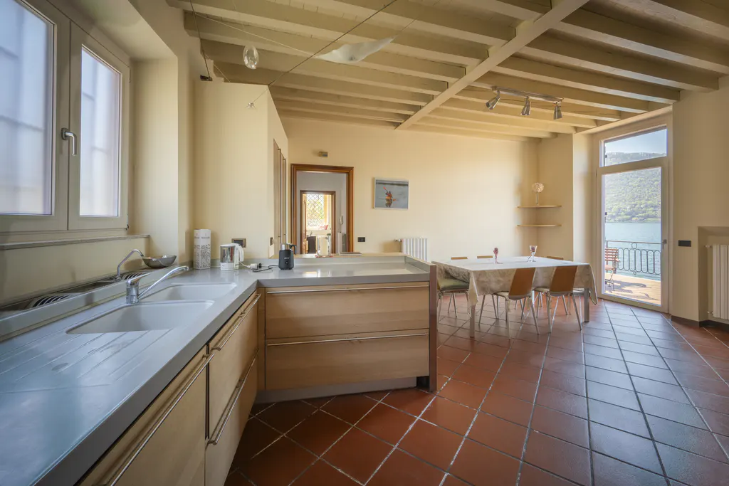 A bright kitchen and dining area with terracotta tile flooring. A sink and cabinets are on the left, and a table with chairs is on the right, with a balcony and lake view in the background.