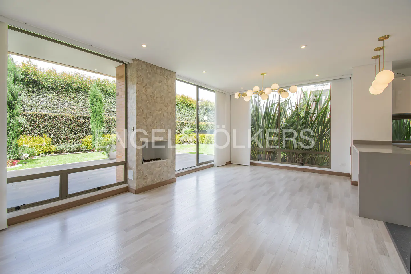 Bright, modern living room with light wood floors, a central fireplace, and large windows overlooking a lush garden.