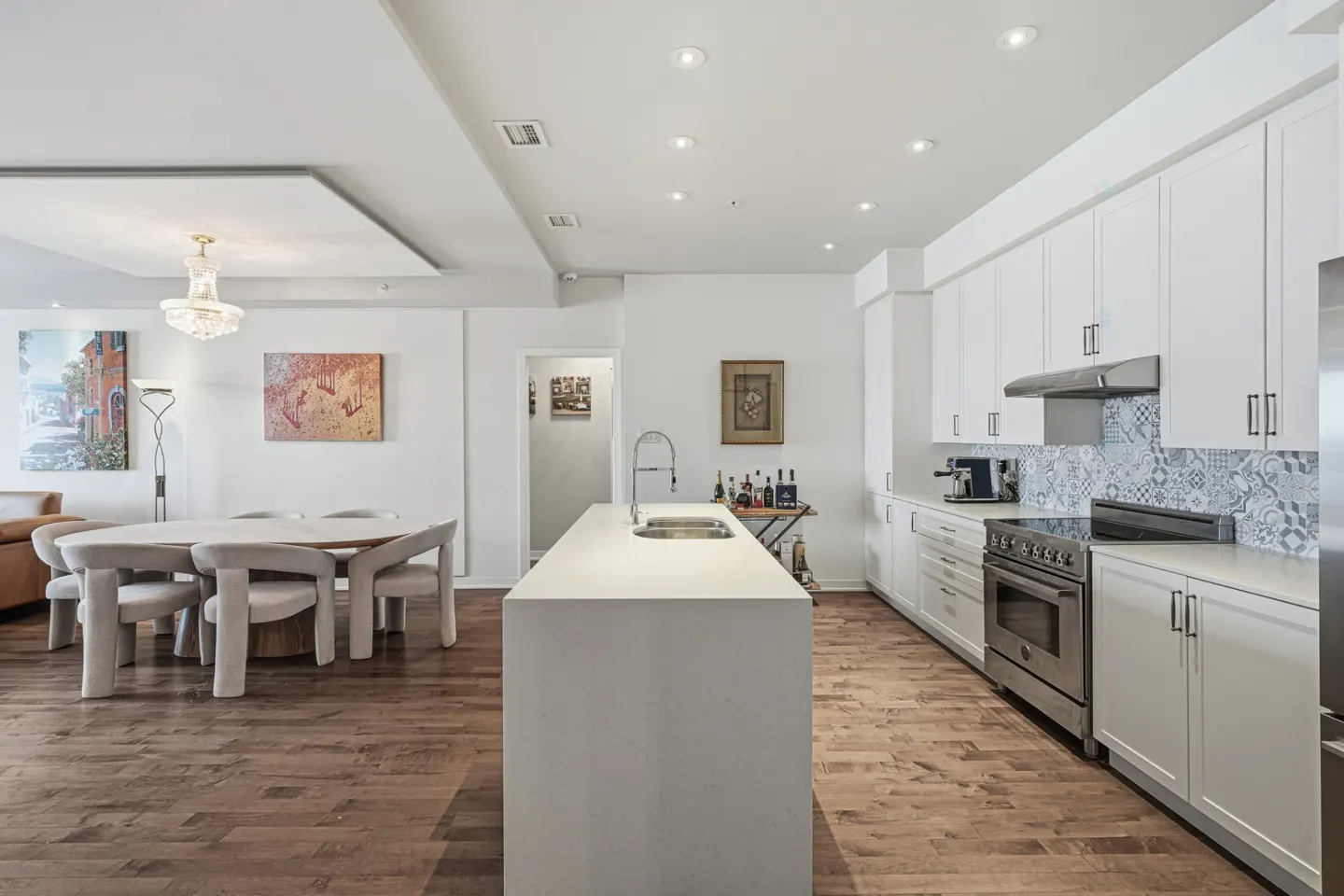 Bright, modern kitchen with white cabinets, island, and stainless steel appliances. Dining area with round table and chairs visible. Hardwood floors throughout.