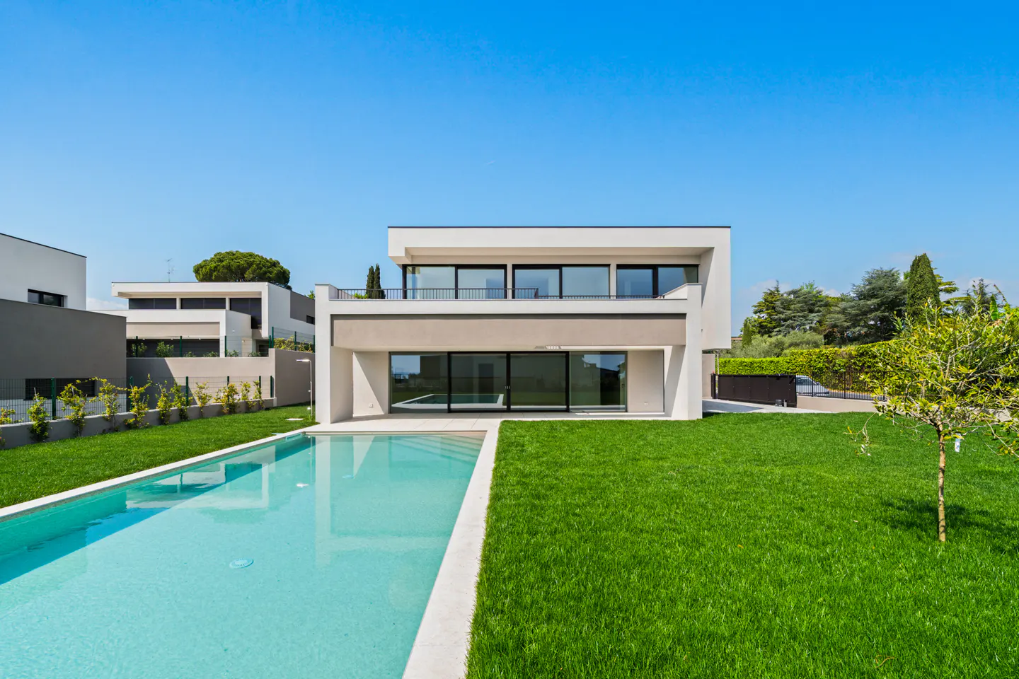 Modern two-story white house with a pool and green lawn under a clear blue sky.