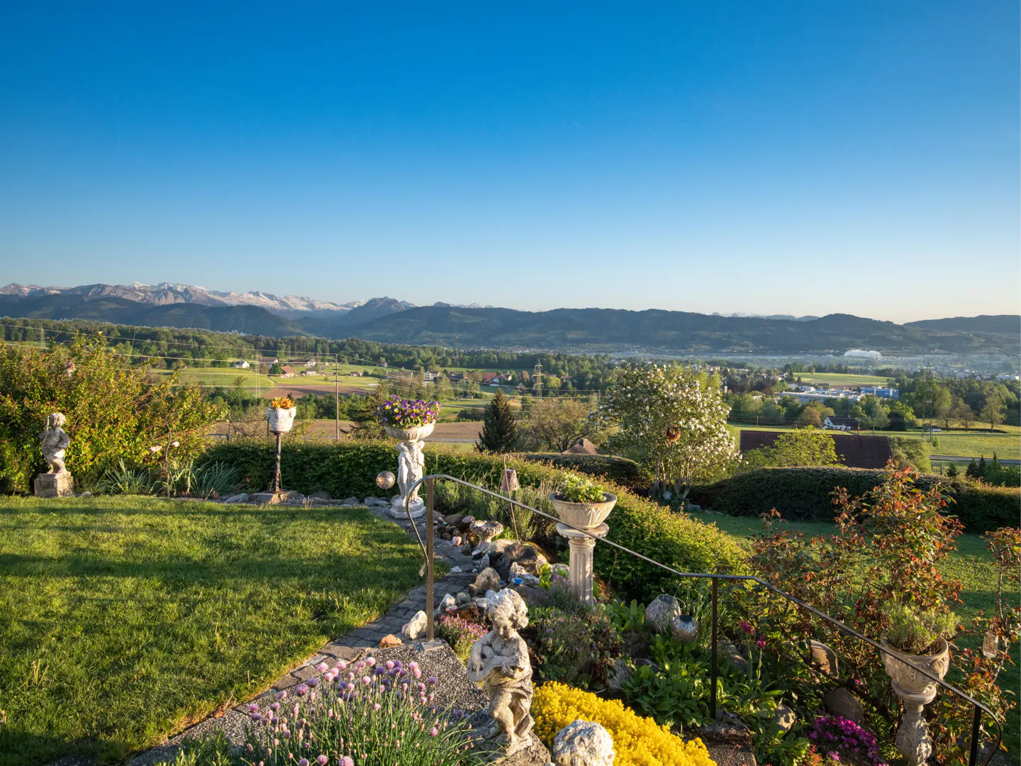 A garden with stone statues and flower pots overlooks a lush valley with distant snow-capped mountains under a blue sky.
