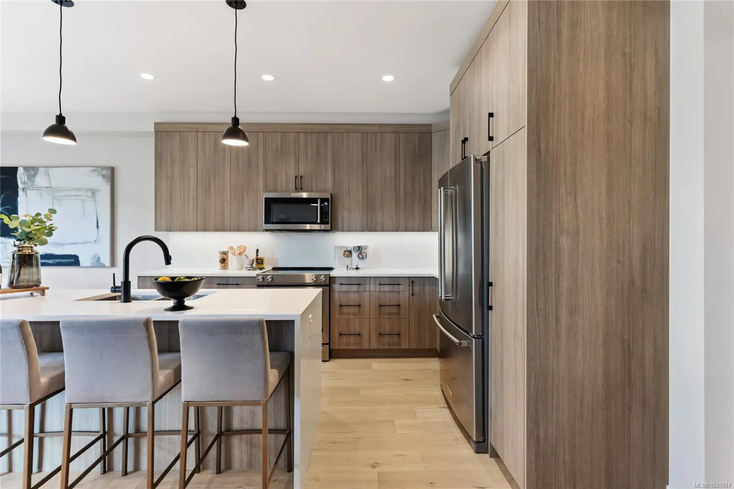 A modern kitchen with wood cabinets, stainless steel appliances, a white countertop island with stools, and black pendant lights.