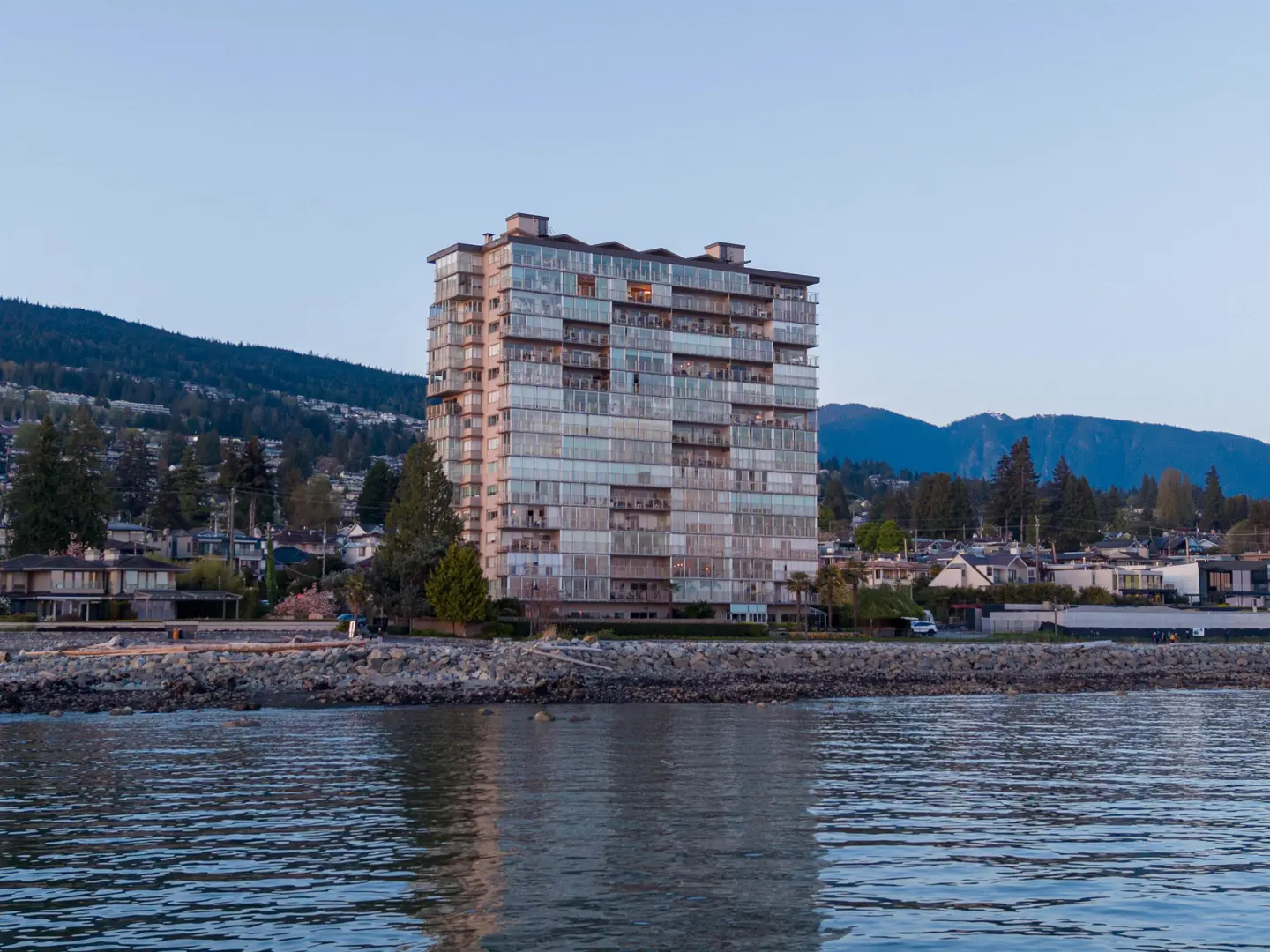 View of a tall, light-colored condo building with many windows, near a rocky shoreline and calm water. Mountains and trees are in the background.