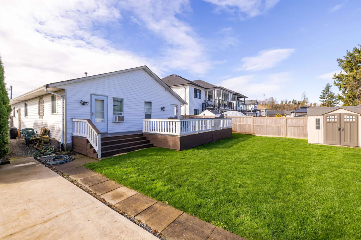 Backyard view of a white house with a brown deck, green lawn, wooden fence, and a shed under a blue sky.