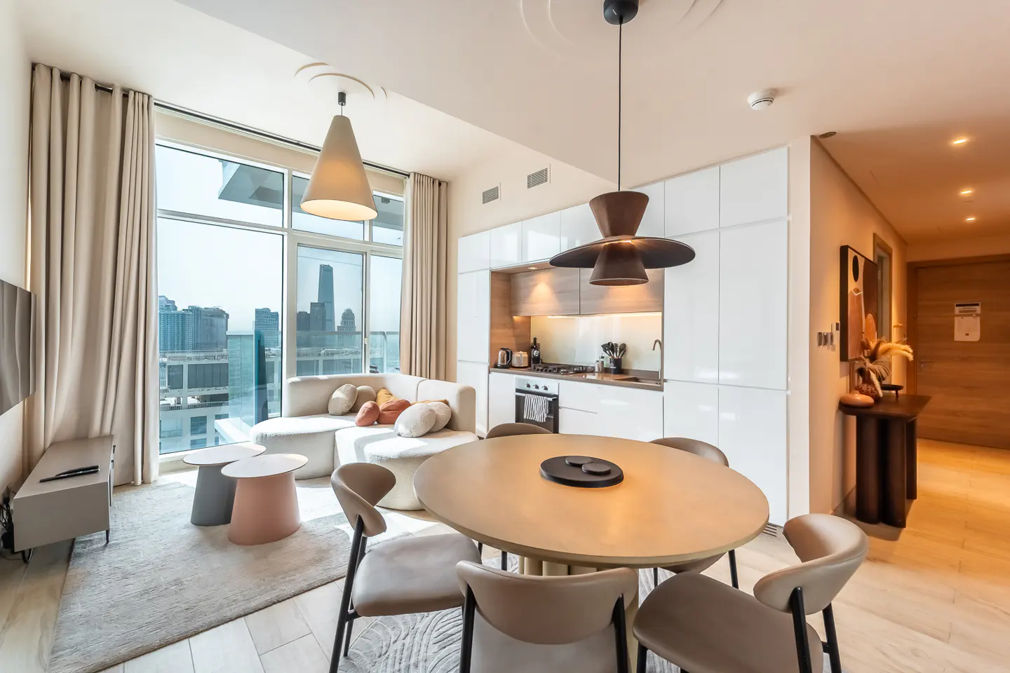 Bright apartment interior with a round dining table, white kitchen, and a sectional sofa by a large window with a city view.