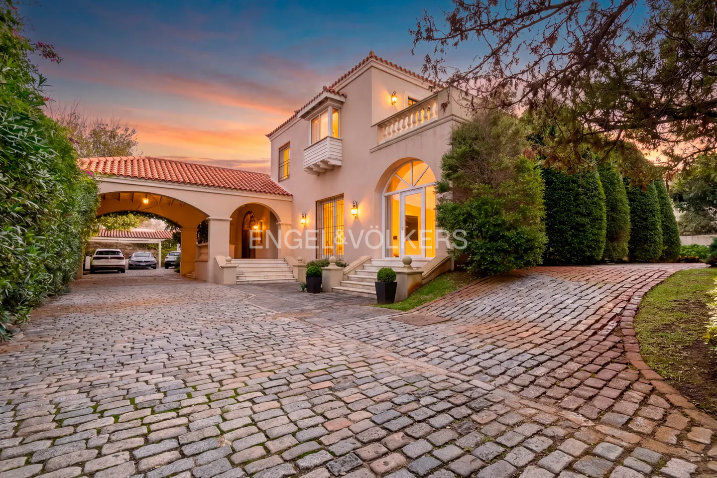 A tan two-story house with a red tile roof and a cobblestone driveway at sunset.