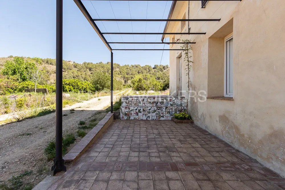 Exterior view of a patio with brick flooring, a tiled bench, and a metal pergola. A hillside with trees is in the background.