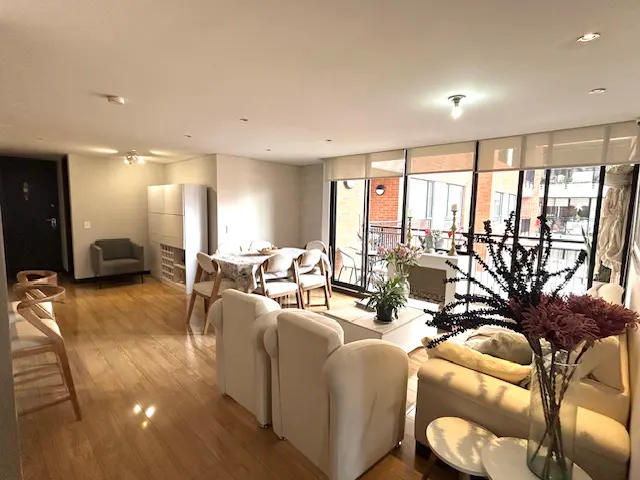 Bright living room with hardwood floors, white sofas, dining table, and a balcony with a view.