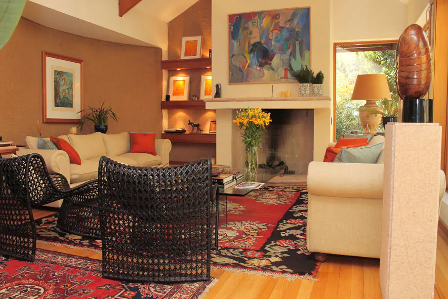 Living room with beige sofas, black wicker chairs, and a red patterned rug. A fireplace and artwork add warmth to the space.