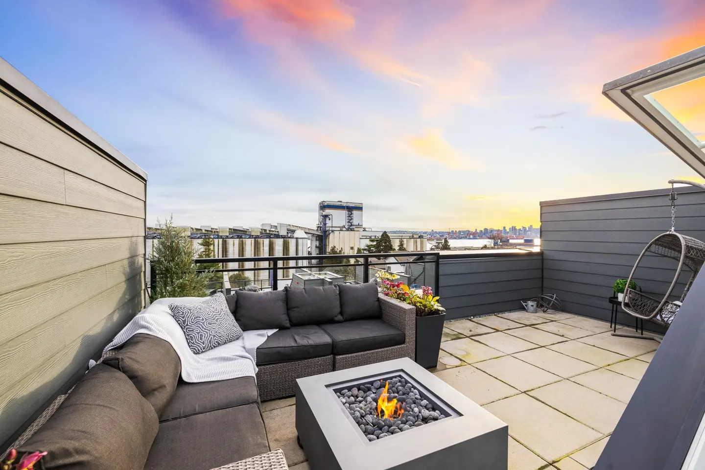 Rooftop patio with gray wicker sofa, fire pit, and hanging chair. City skyline and colorful sunset in the background.