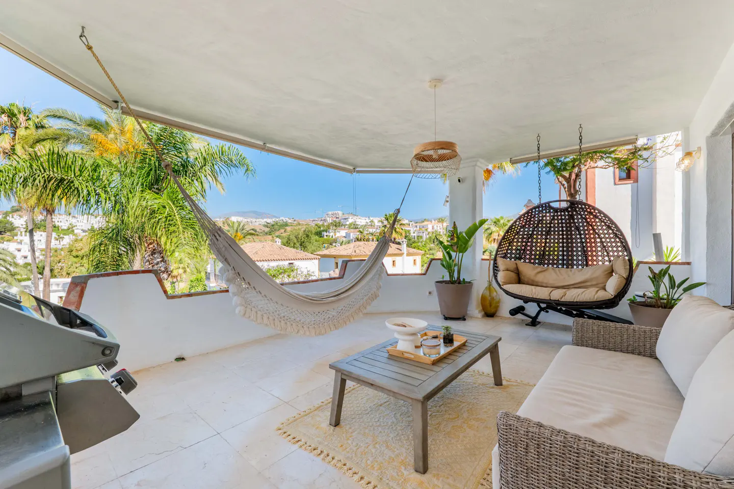 Covered terrace with a hammock, hanging egg chair, sofa, coffee table, and scenic hillside view under a clear blue sky.