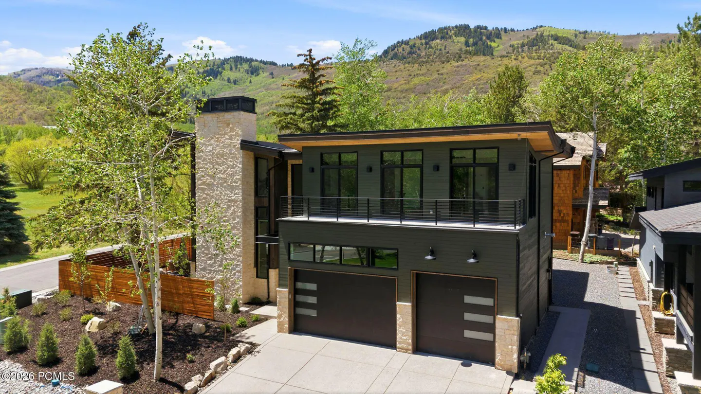 Modern two-story home with a balcony, dark siding, and a stone chimney. Mountains and trees are in the background.