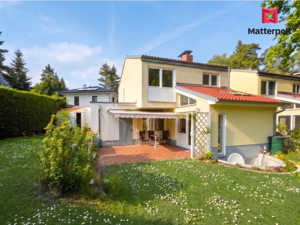Backyard view of a yellow townhouse with a red tile roof, a brick patio, and a green lawn with white flowers.