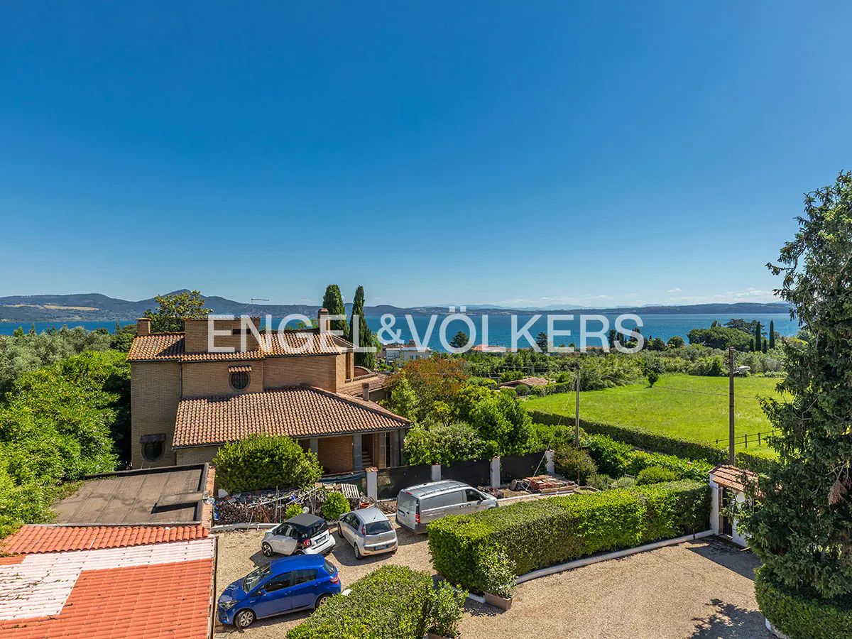 Real estate view of a tan house with a red tile roof, cars in the driveway, and a lake in the background under a blue sky.