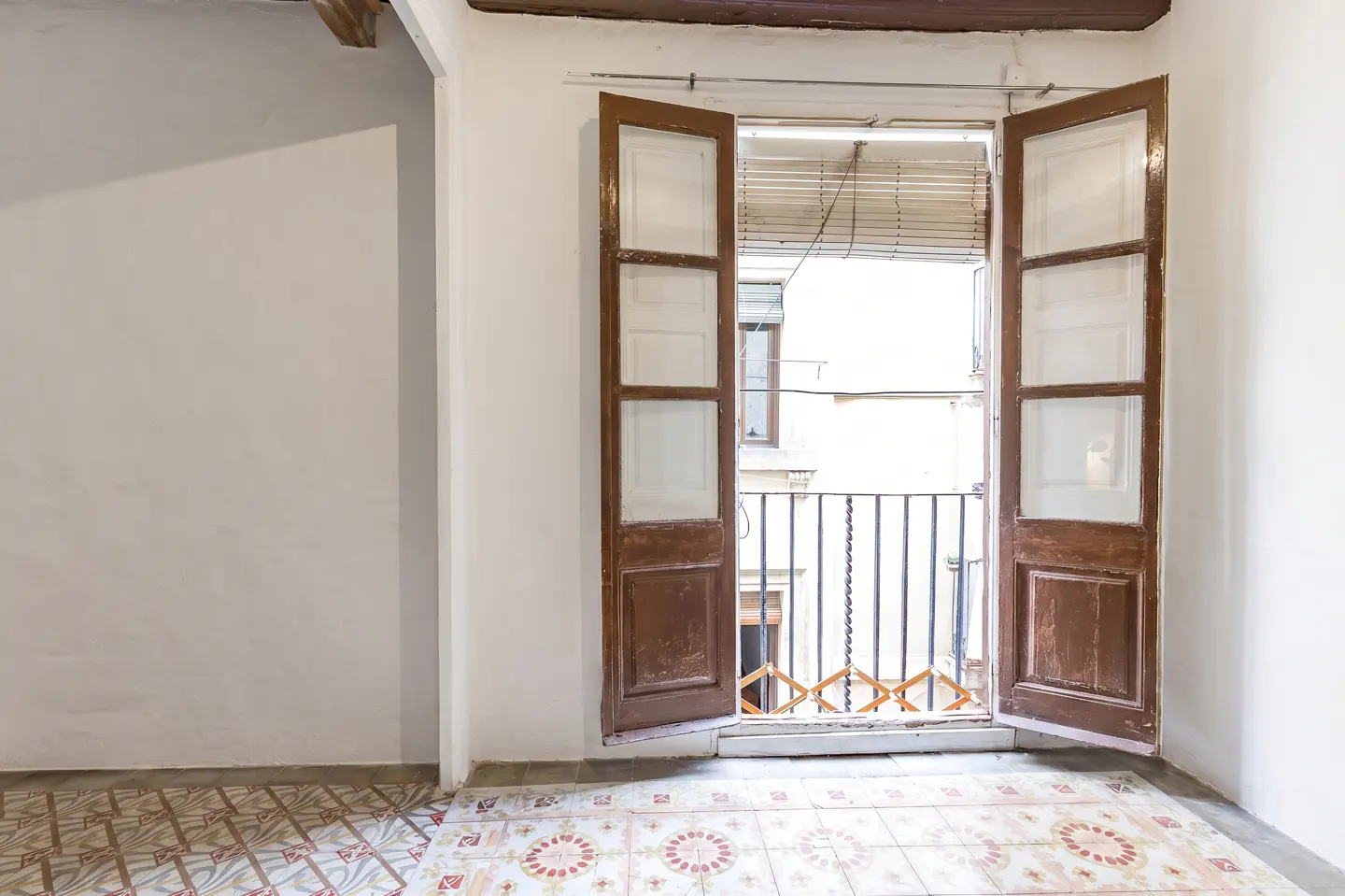 A bright room with white walls and patterned tile floor, featuring open brown doors leading to a balcony with a black railing.