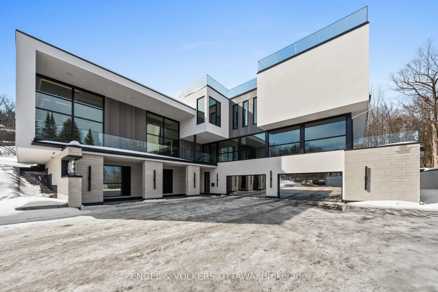 Modern white house with large windows and glass railings. The driveway is paved and partially covered in snow. Blue sky.