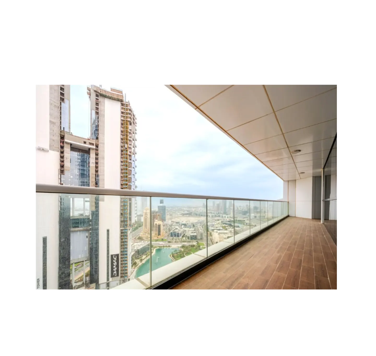 Balcony view of Dubai skyline with glass railing, brown tiled floor, and modern ceiling. Tall buildings and a blue lake are visible in the distance.