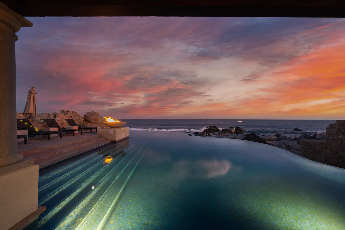 Infinity pool at sunset with a fire pit, lounge chairs, and ocean view under a vibrant pink and orange sky.