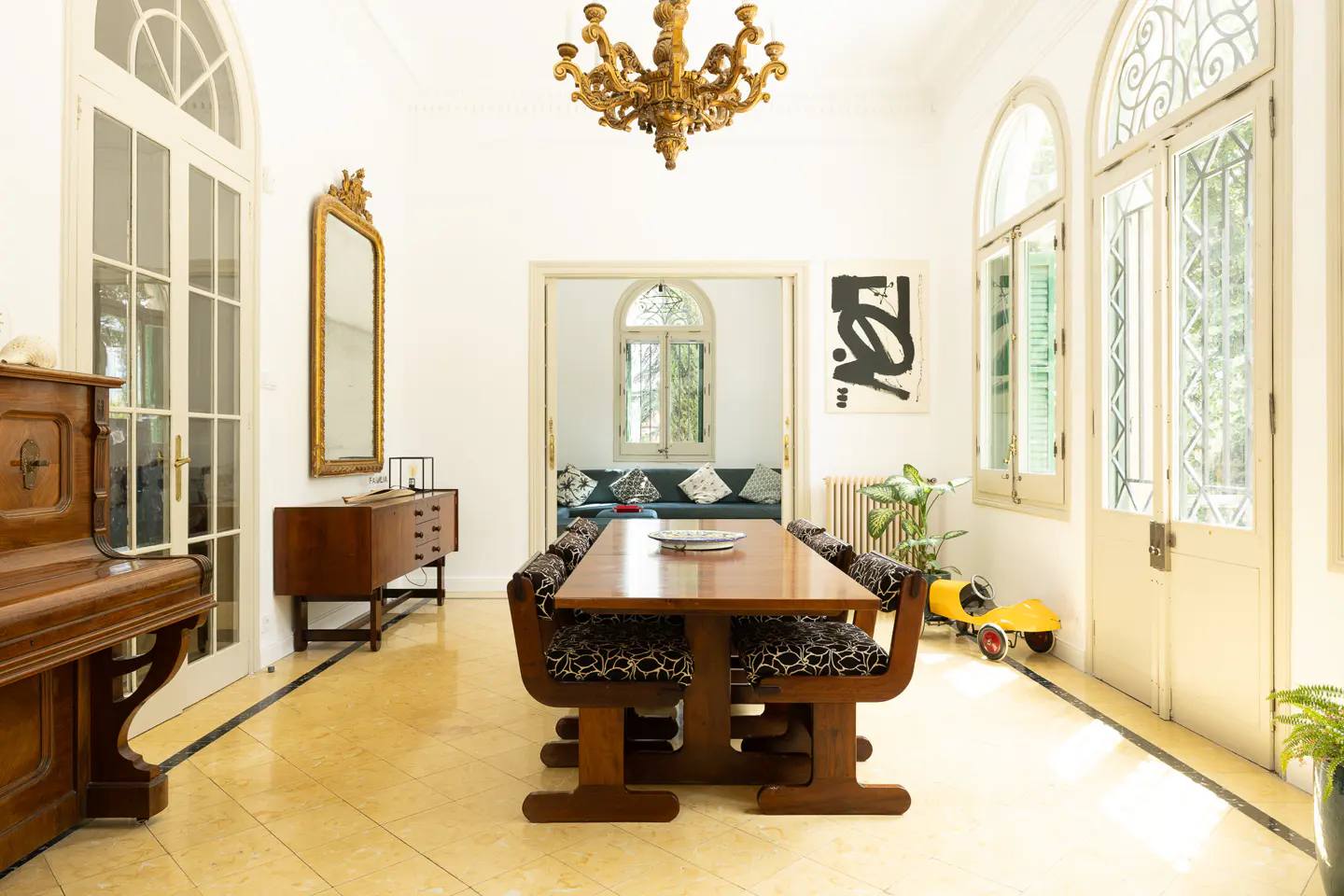 Bright dining room with a long wooden table, patterned chairs, and a golden chandelier. A piano and vintage cabinet are on the left. Arched windows let in light.