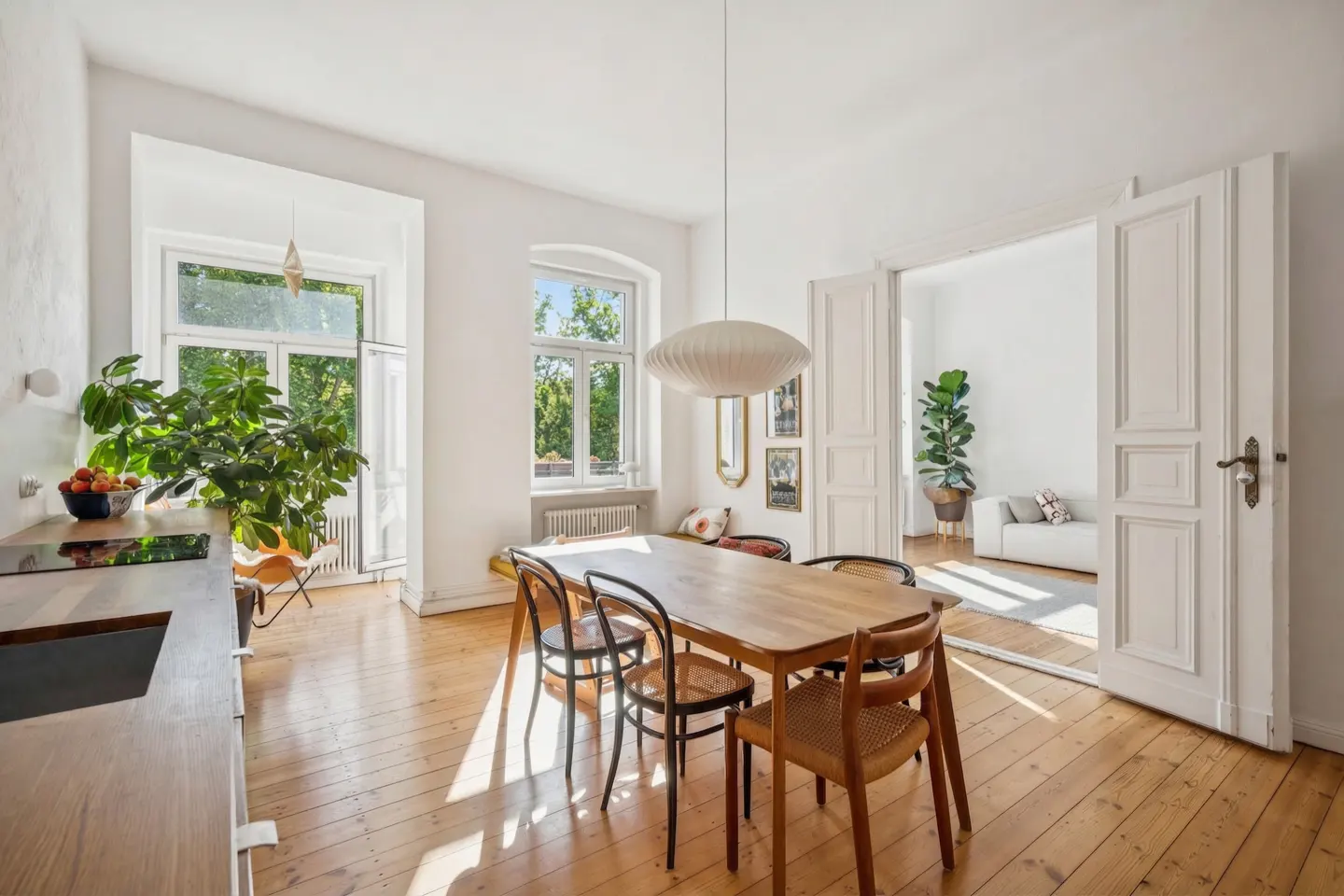 Bright dining room with wood floors, table, and chairs. White walls, windows with green trees outside, and open doorway to living room.