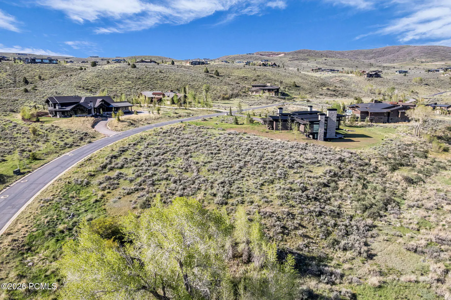 Aerial view of luxury homes on a grassy hillside under a blue sky with scattered clouds. A paved road winds through the landscape.