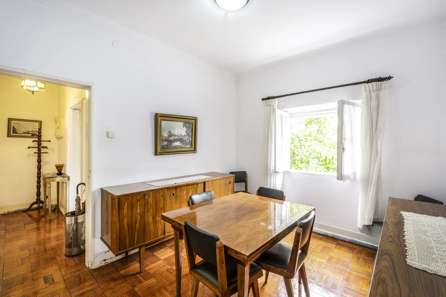 Bright dining room with wood floors, table, and chairs. A wooden cabinet sits against the wall with a framed picture above it. Natural light streams through a window.