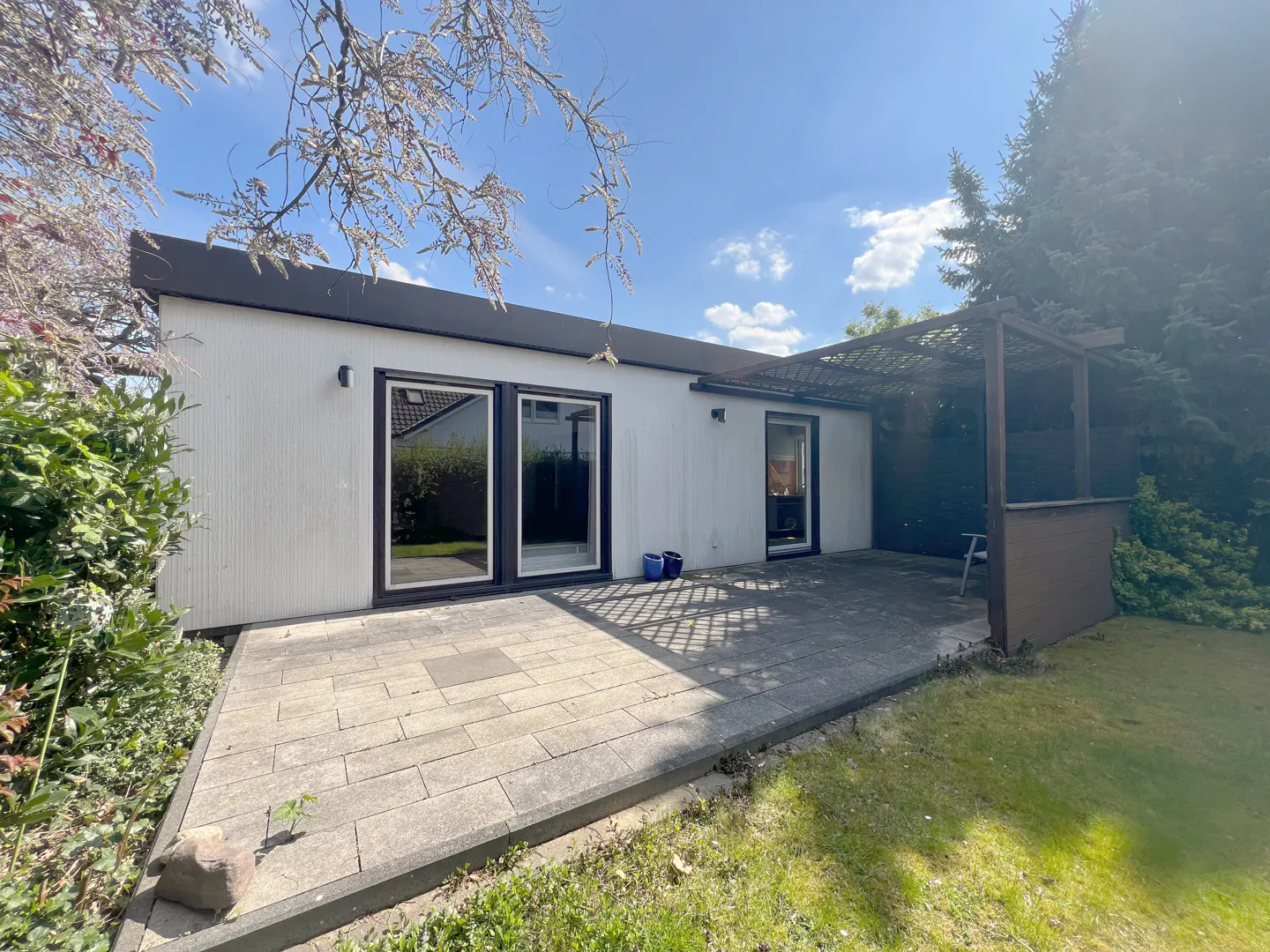 Exterior view of a white, modern home with a patio, pergola, and green lawn on a sunny day.