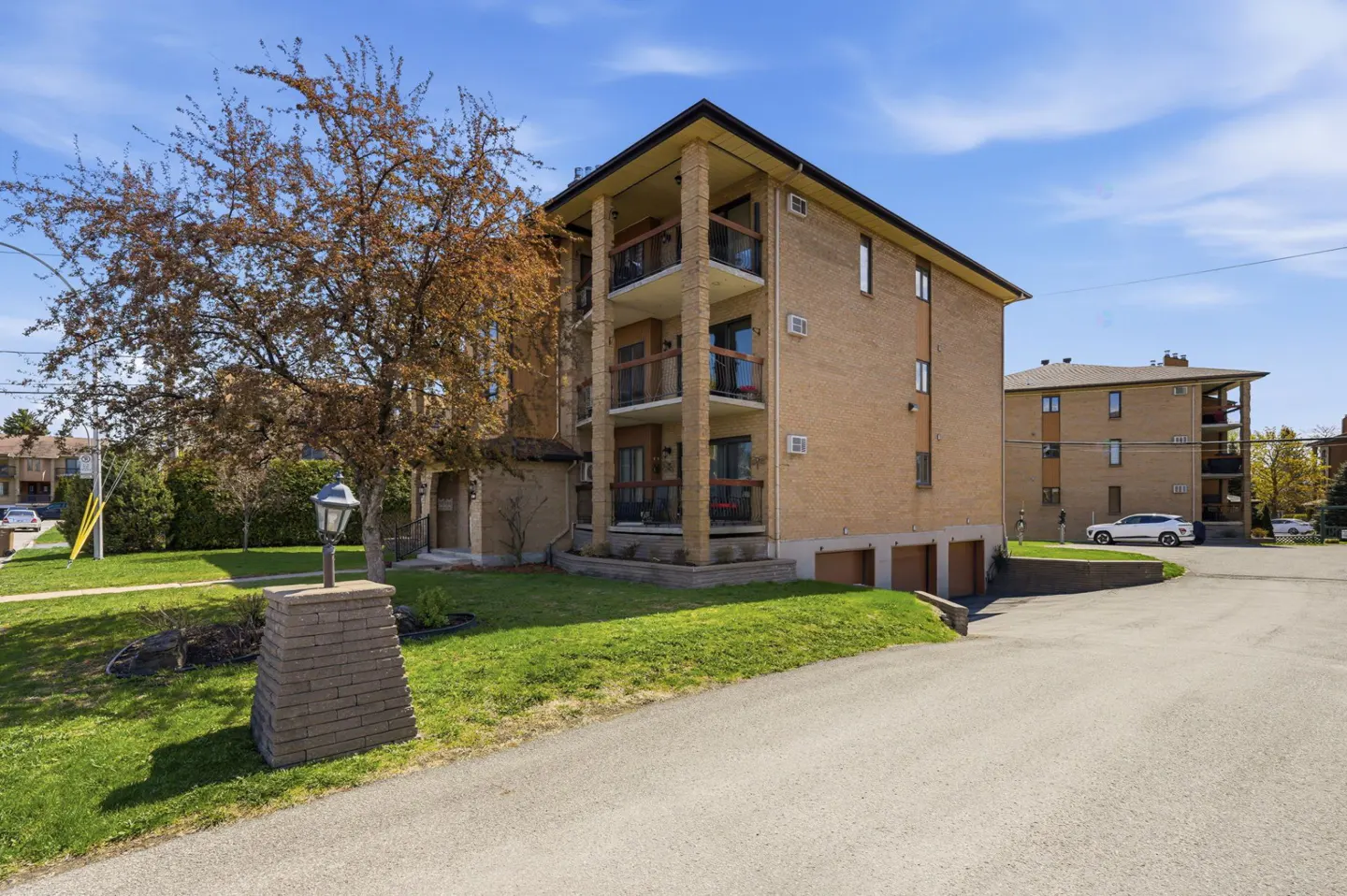 A three-story brick apartment building with balconies, green lawn, and a tree under a blue sky.