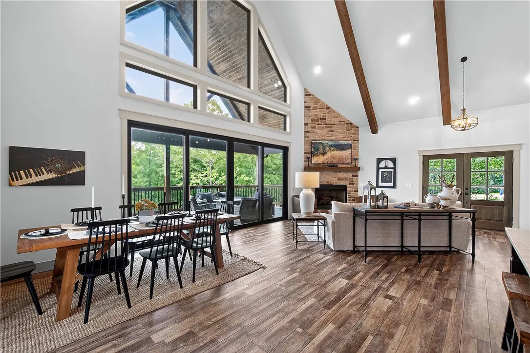 Open-concept living area with wood floors, vaulted ceilings, stone fireplace, dining table, and large windows to a deck.