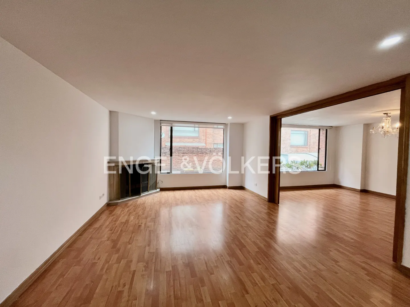 Bright, empty living room with hardwood floors, white walls, and a fireplace. Large windows offer natural light. An open doorway leads to another room.