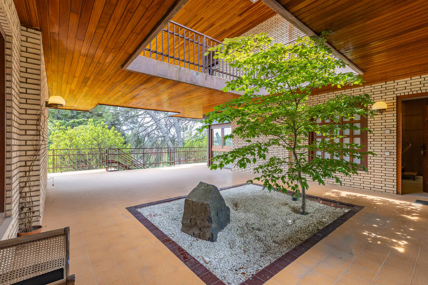 Modern courtyard with a central tree, a large stone, and gravel, set under a wooden ceiling with a brick wall backdrop.