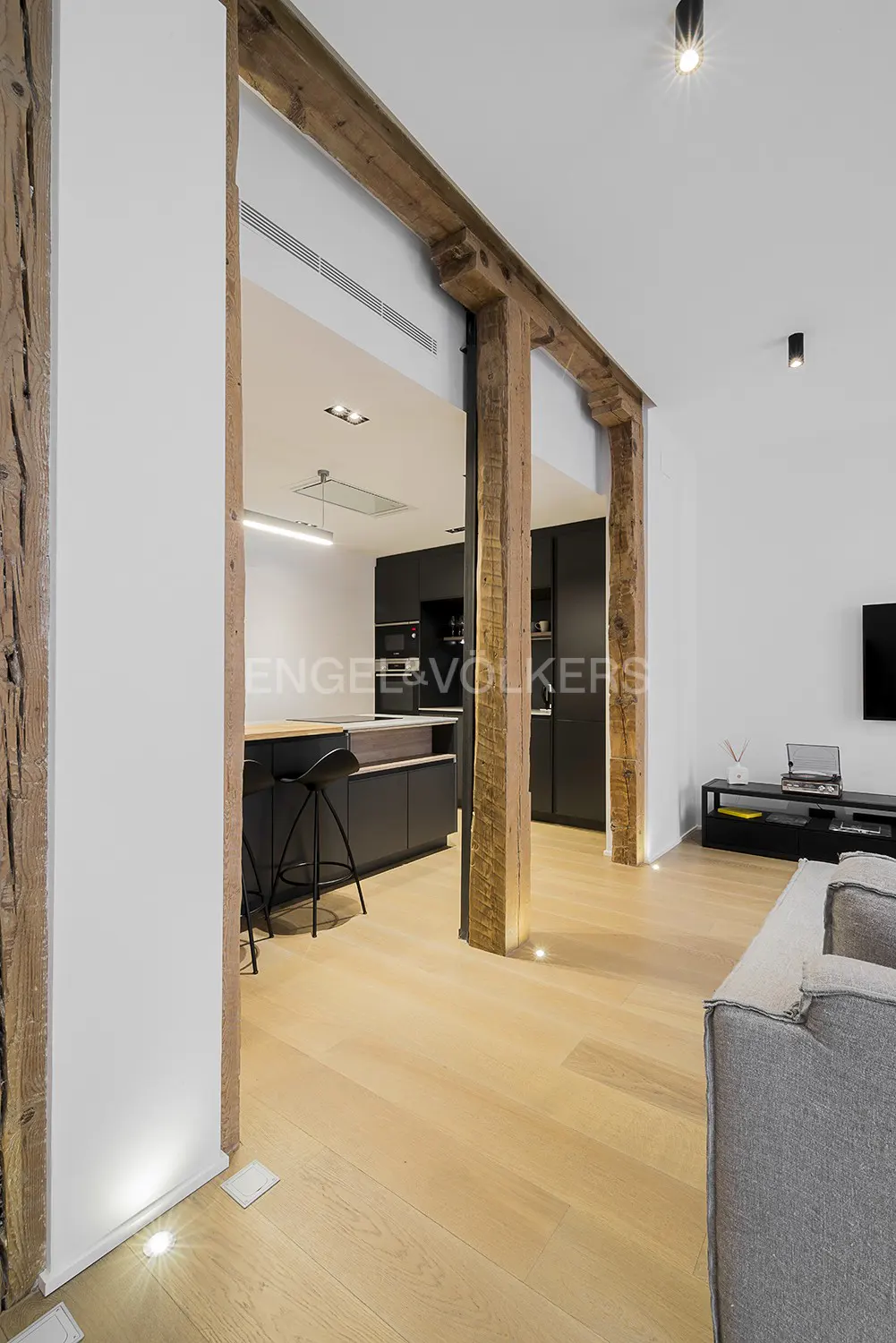 Open-concept living space with wooden beams separating the kitchen. Black cabinets, island with stools, and light wood floors.
