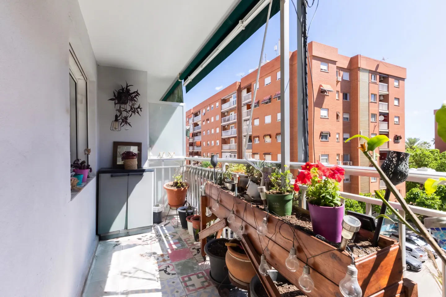 A sunlit balcony with potted plants and string lights, overlooking a red brick apartment building.