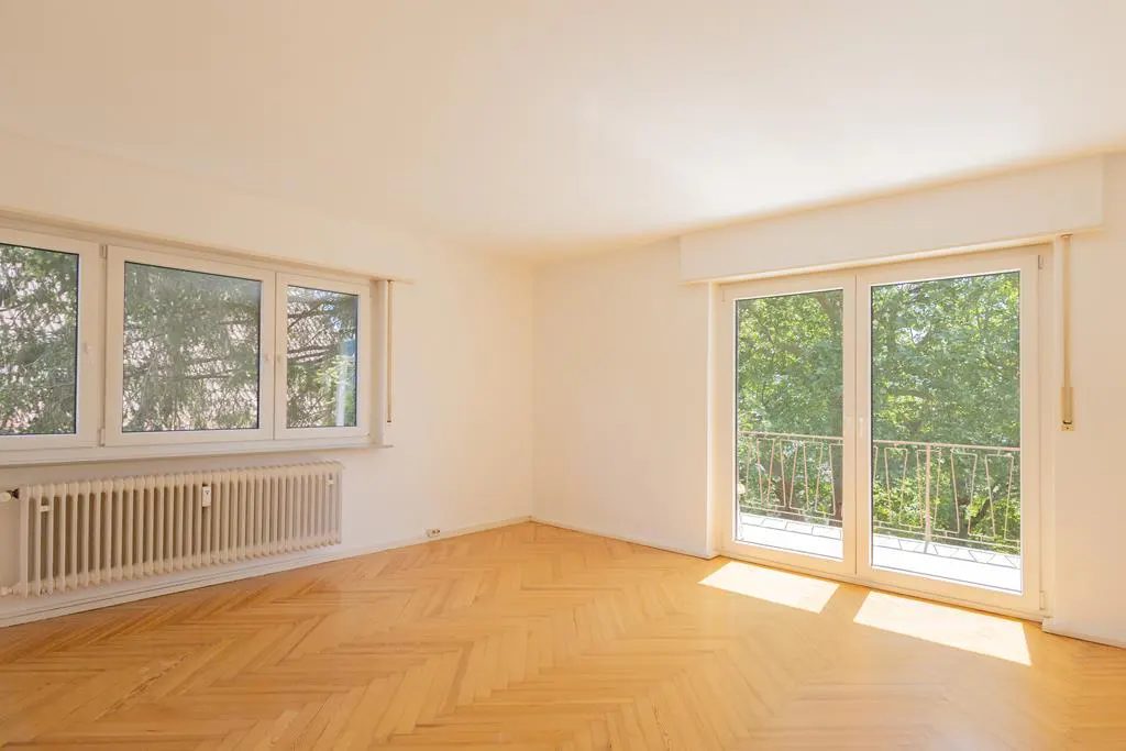 Bright, empty room with herringbone wood floors, white walls, and a radiator under a window. Balcony doors show green trees outside.