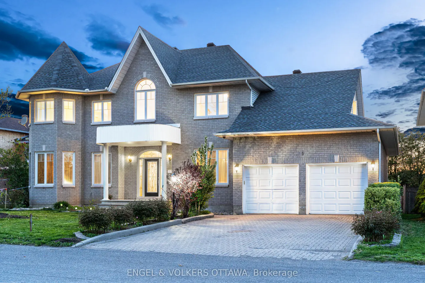 Two-story gray brick house with a turret, white garage doors, and a brick driveway at dusk.