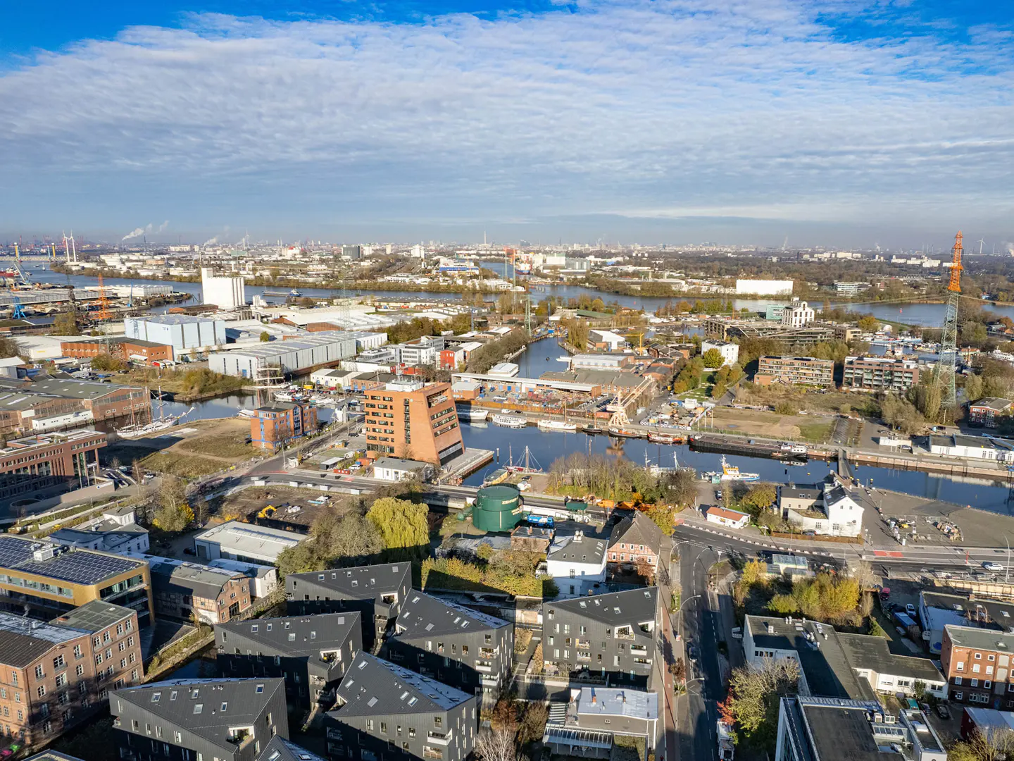 Aerial view of Hamburg, Germany, featuring modern black-roofed buildings, waterways, and industrial areas under a partly cloudy sky.