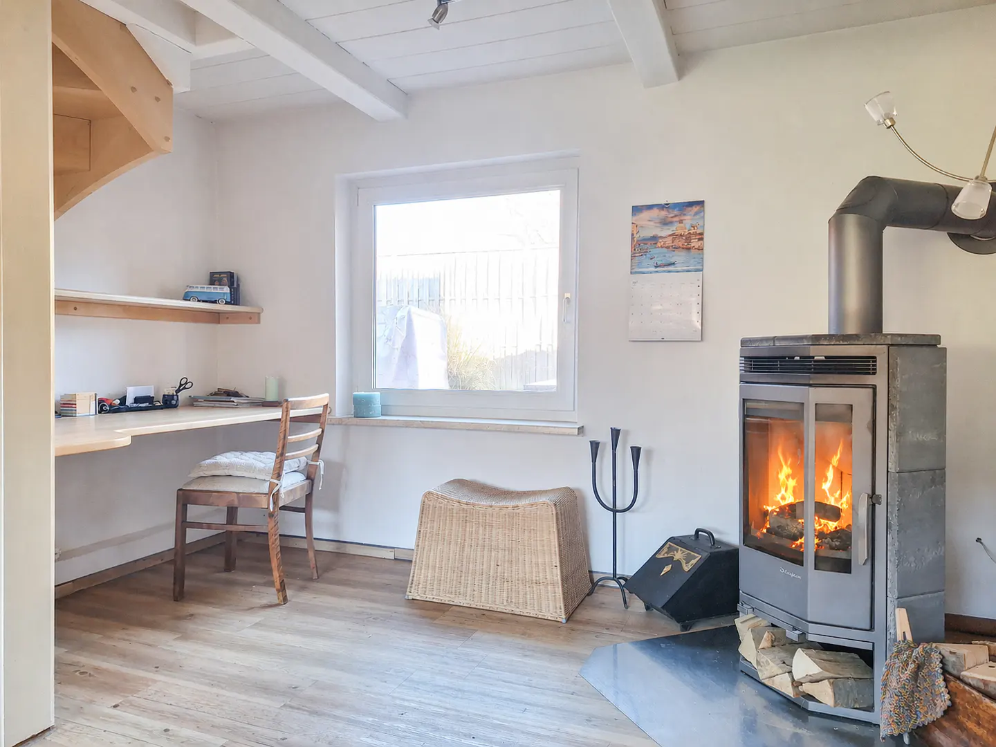 Bright room with a wood-burning stove, desk, and window. The stove is gray with a glass door showing flames. A wooden desk and chair are to the left.