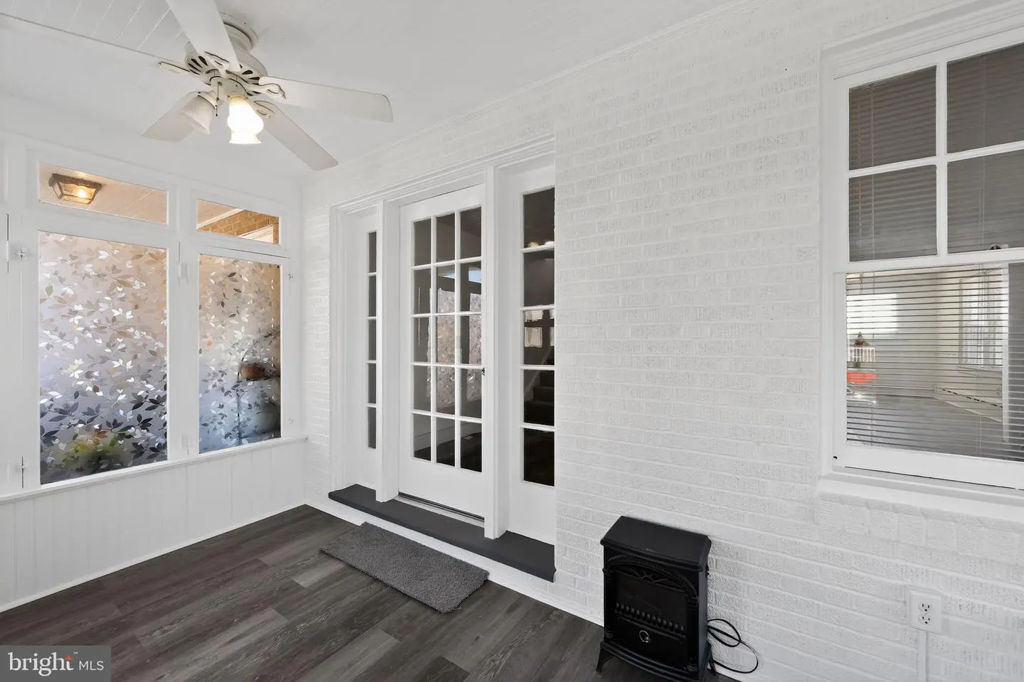 Bright sunroom with white brick walls, dark wood floors, and a white ceiling fan. Windows have frosted leaf patterns. French doors lead outside.