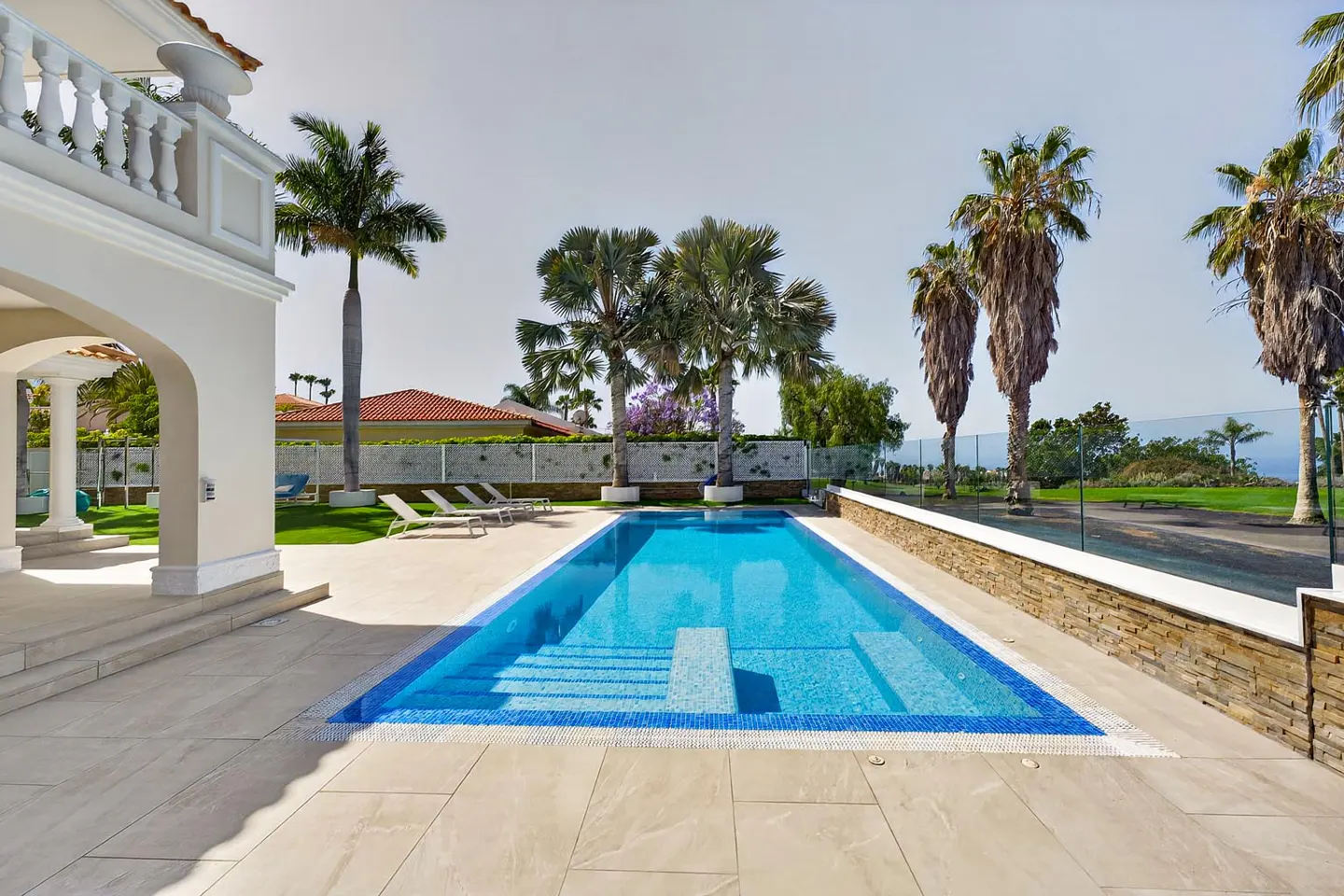 A rectangular blue pool with steps, surrounded by palm trees and a white building with arches.